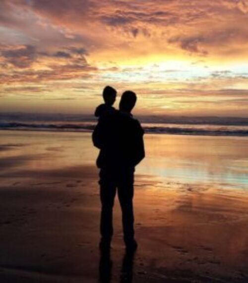 Silhouette of an adult holding a child while watching a colorful sunset over the ocean.