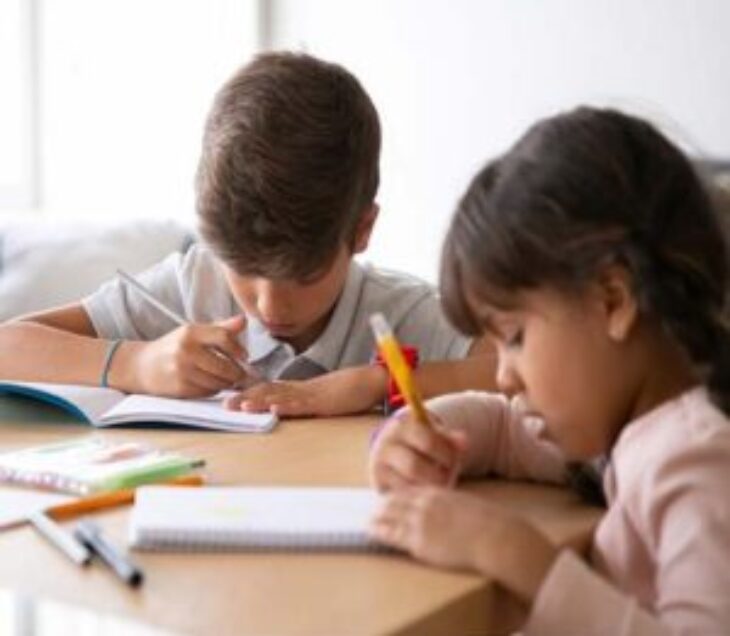 Two young children sitting at a table, focused on writing and drawing in their notebooks.